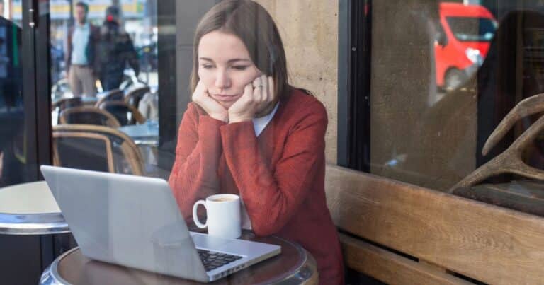 Woman in a red sweater looks contemplative while seated at a cafe with her laptop and a cup of coffee.