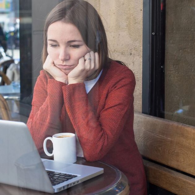 Woman in a red sweater looks contemplative while seated at a cafe with her laptop and a cup of coffee.